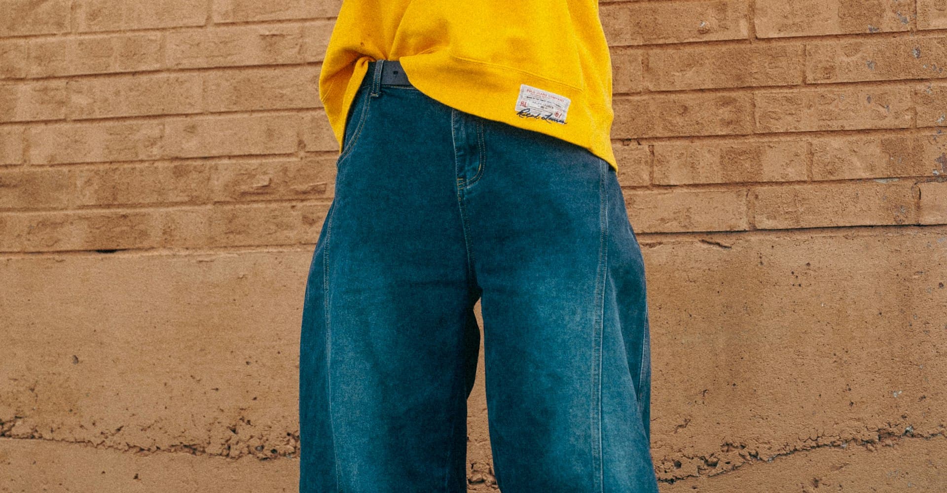Man in a yellow sweatshirt and baggy blue jeans against a brick wall